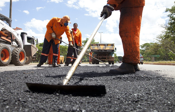 No canto esquerdo da avenida dois caminhões estão estacionados e três Homens com uniformes amarelos empunham pás e fazem o serviço de tapa-buraco