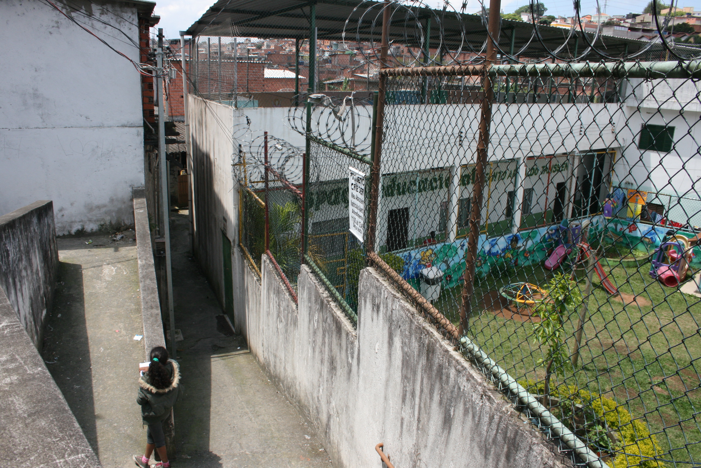 Imagem mostra um gramado com brinquedos infantis de um centro educacional à direita, com uma criança encostada em uma mureta ao centro da imagem, do lado de fora do centro educacional