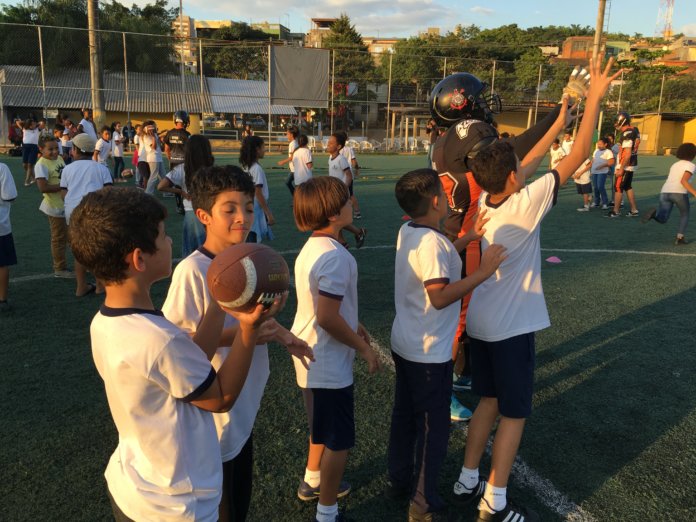 Com orientação do atleta crianças no gramado lançam a bola americana