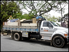 Caminhão da operação Cata-Bagulho com a caçamba lotada de pedaços de madeira e móveis velhos. Em cima da caçamba também é possível ver um funcionário da Subprefeitura Ipiranga colocando entulho dentro do caminhão.