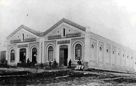 Foto antiga, em preto e branco, mostra a Fábrica de Chocolates Falchi, a primeira indústria de Vila Prudente. A foto data do ano de 1897, e traz algumas pessoas em frente ao prédio, que é térreo, com duas entradas e que se estende até o fundo da fotografia.