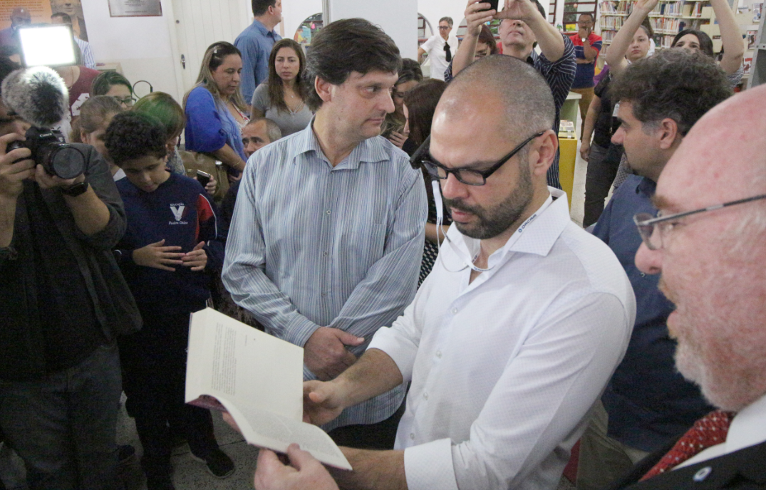 O prefeito Bruno Covas testando o óculos. Ele está com um livro nas mãos.
