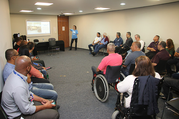 assessora técnica da SMPED Carolina Santos durante apresentação, dentro de uma sala. As pessoas observam ao redor. Ela aponta para um telão que está atrás dela, com uma apresentação.