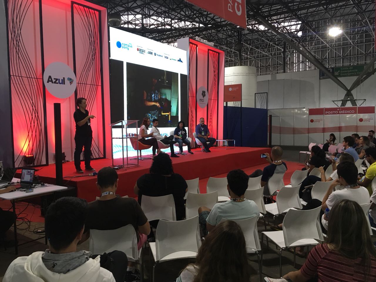 mediadora e palestrantes no palco. Atrás, um telão com imagens. Lado direito, a intérprete de Libras. Público observa a palestra. Todos estão sentados em cadeiras, posicionadas de frente para o palco.