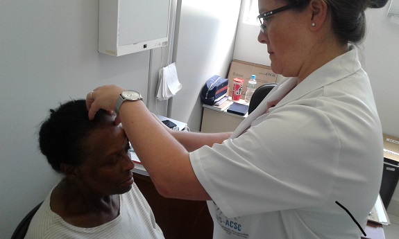 Em uma sala branca, a médica de jaleco branco, examina a cabeça da paciente. Ao fundo, uma mesa branca e uma janela com cortinas fechadas.