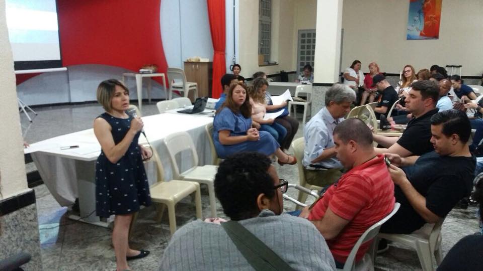  a secretária adjunta da SMPED, Marinalva Cruz, conversa com os participantes. Ela segura um microfone nas mãos. A presidente do CMPED e demais conselheiros estão sentados. 
