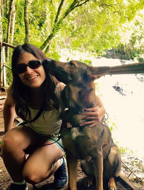 Marina com o seu cão guia Duke. Ela está encurvada, com o cão ao seu lado e abraça ele. Ele é um pastor alemão. Ela sorri, usa óculos escuros e roupa esportiva. 
