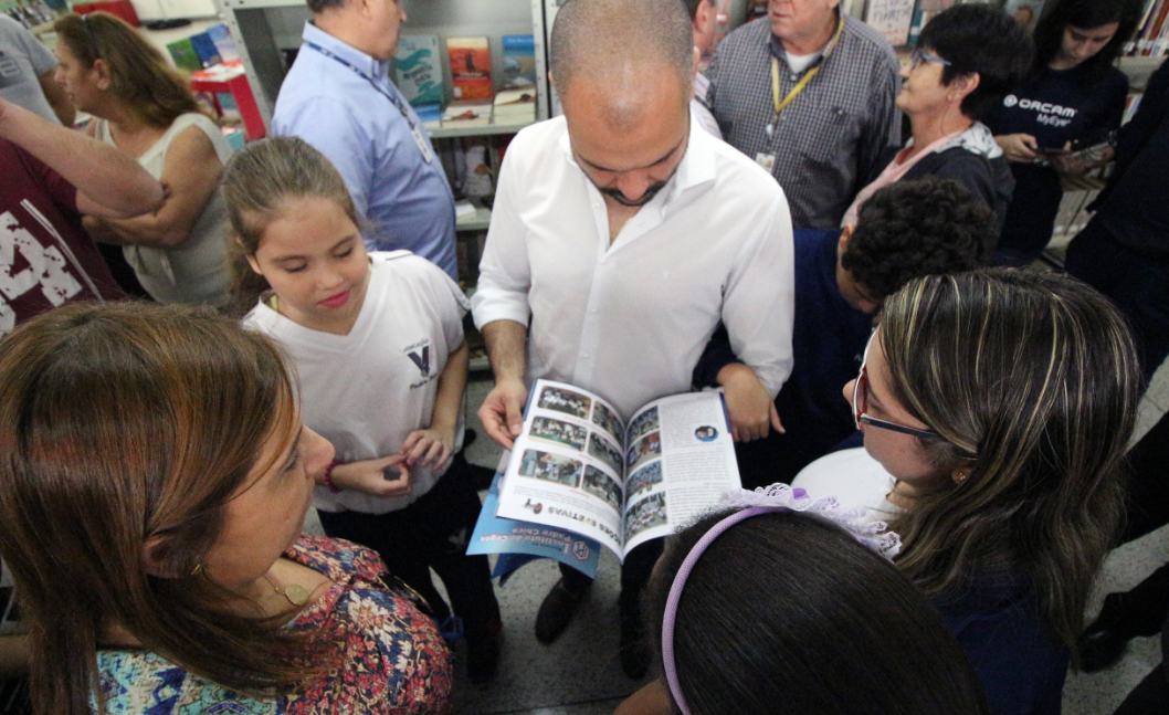 O prefeito Bruno Covas conversa com as crianças e segura um livro nas mãos.