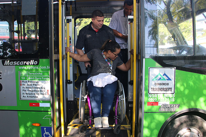 a arquiteta cadeirante Myrna, da SMPED, participa da viviência em um ônibus. Um homem ajuda ela descer do ônibus, segurando atrás de sua cadeira de rodas.