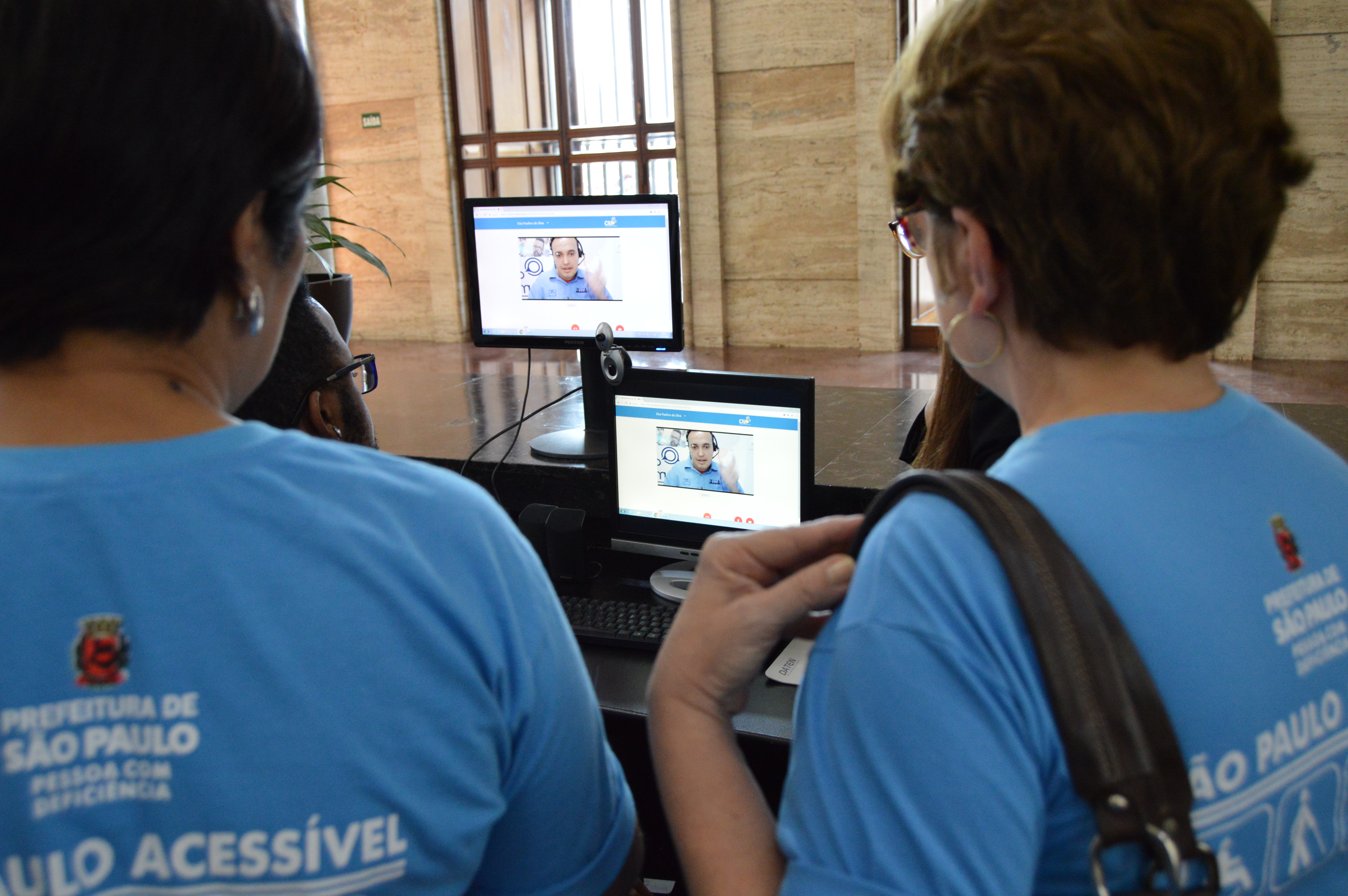 Intérprete de Libras na tela do computador, por meio do aplicatico da CIL, conversa com a equipe da Prefeitura de São Paulo, que está salão de entrada da Prefeitura de São Paulo.