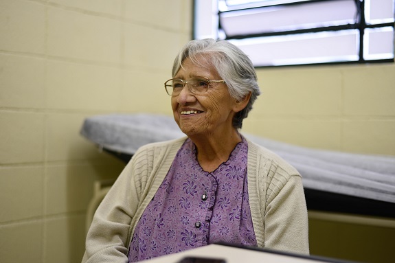 Dona Olga de óculos e sorrindo está sentada no consultório. Veste uma camisa lilás e blusa creme.Ao fundo uma maca e uma janela.