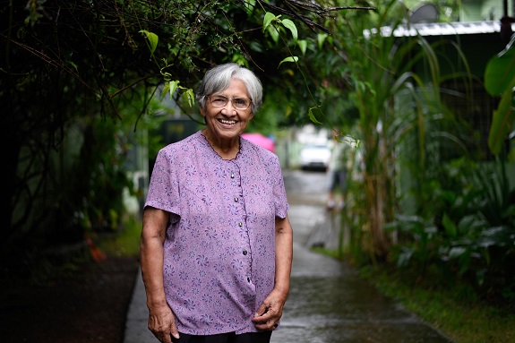 Dona Olga com uma camisa lilás em um corredor externo arborizado