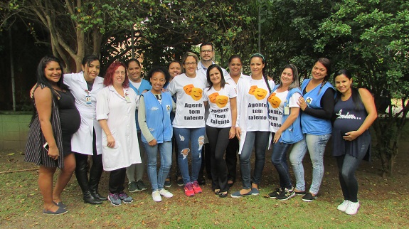 Grupo posa em um jardim. Entre eles, duas gestantes, profissionais de jaleco branco, agentes de saúde de colete azul e pessoas com camisetas personalizadas escrito: Agente tem talento.