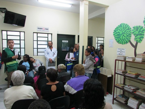 Na sala de espera iluminada, pacientes sentadas ouvem o agente de saúde. À direita, uma estante de madeira com livros e a ilustração de uma árvore na parede.