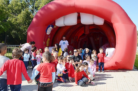 Crianças brincam em área externa. Ao fundo, um túnel gigante enchido com ar em forma de uma boca com dentes para representar do caminho da voz.