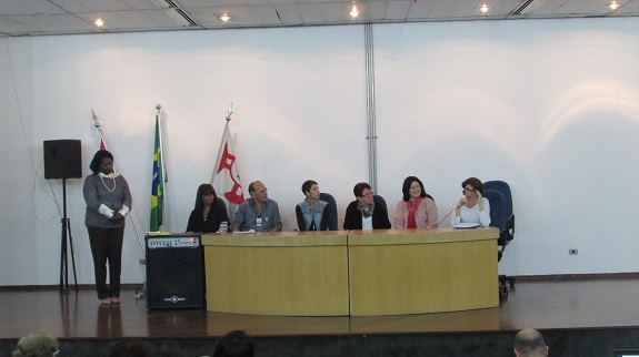 Em um palco, convidados sentam atrás de uma mesa de madeira marfim. À esquerda, bandeiras, uma caixa de som e uma participante em pé. Ao fundo, uma prede branca.