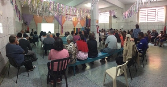 Em uma sala grande com paredes de azulejos, participantes estão sentados em bancos organizados em fileiras. No teto há enfeites roxos e laranjas pendurados por todo o salão.