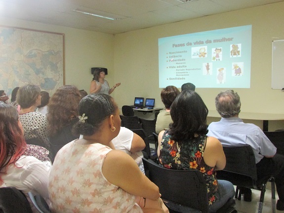 Participantes assistem à palestra em sala de reunões. Ao fundo, uma tela projetando as fases da vida de uma mulher.