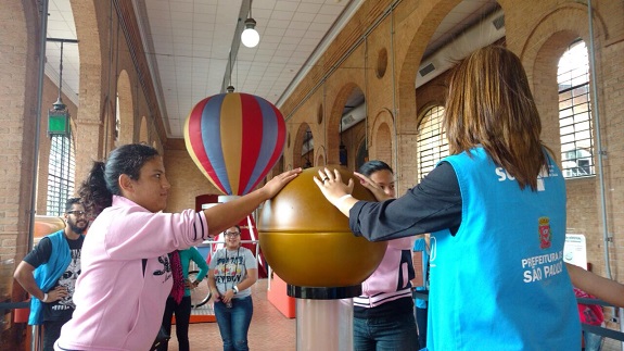 Educadora, de colete azul com logotipo da prefeitura de São Paulo, e usuária, de moletom rosa claro,seguram as duas mãos em uma bola dourada. Ao fundo, um balão listrado de amarelo, vermelho e azul.