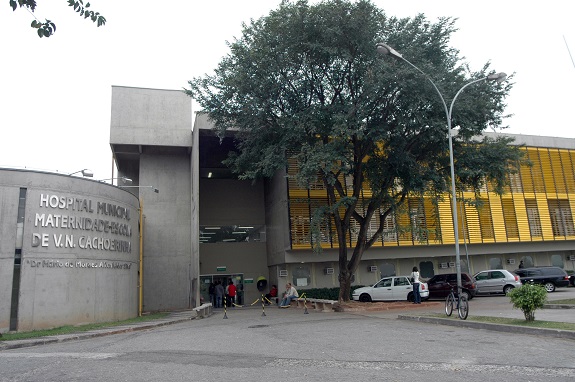 Fachada do Hospital Municipal Maternidade Escola de Vila Nova Cachoeirinha em cinza e prata.. Ao centro uma árvore, à direita carros no estacionamento, e ao fundo, o prédio com janelas amarelas.