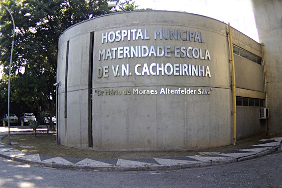 Fachada de cimento do Hospital Municipal Maternidade - Escola de Vila Nova cachoeirinha. Ao fundo, árvores e alguns carros estacionados.