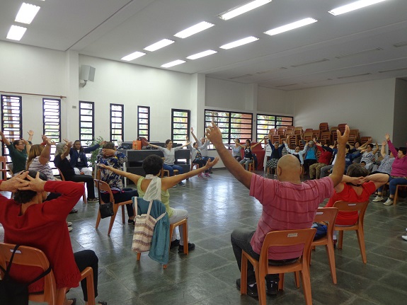 Em uma sala grande com parades brancas, pisos quadriculados e cinza, pessoas com os braçoes erguidos estão sentadas em cadeiras de plástico marrom formando uma roda. Ao fundo, cadeiras empilhadas e janelas de vitrô. 