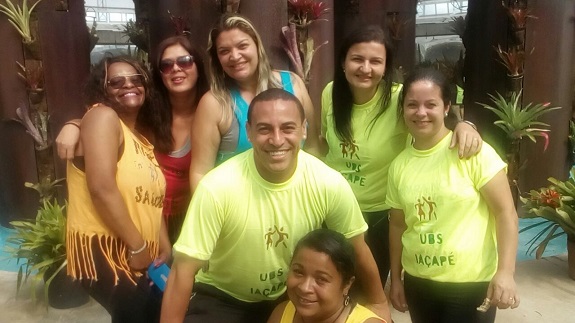 Grupo caracterizado com camisas verde - claras escrito UBS Iaçapé posam em frente a uma casa marrom. O local está enfeitado com plantas ornamentais.