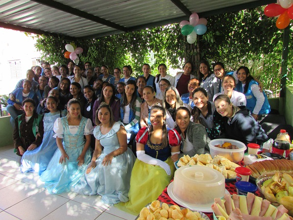 Mulheres posam para a foto. À frente, participantes estão fantasiadas de princesas. À direita, uma mesa farta com pães, frios, bolos, requeijão e refrigerante. Ao fundo, árvores verdes.