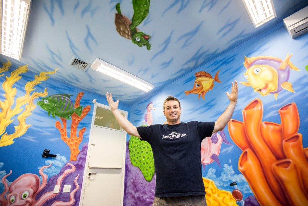 O artista plástico Jason Hulfish posa em frente à sua obra com o tema "fundo do mar", em que desenhou em uma parede algas, peixes, cavalos marinhos e estrelas do mar em vários tons de verde, amarelo, rosa e laranja.