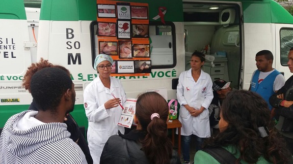 Profissionais de jaleco branco e touca conversam com pacientes em frente à Unidade Móvel. Na janela da unidade móvel, um banner mostra fotos de doenças bucais.