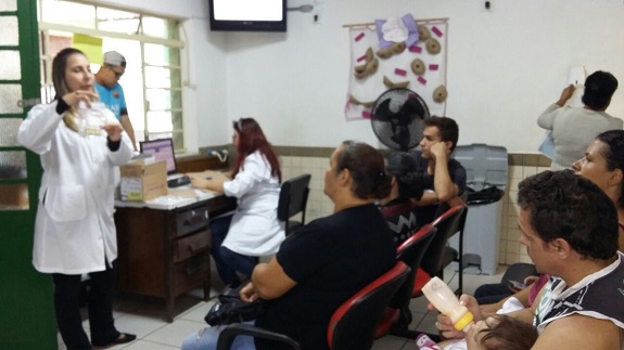 Na sala de espera, profissional de jaleco branco dá instruções a pacientes. Ao fundo, uma janela branca e acima uma televisão de led. Em frente à janela, uma outra profissional sentada, utiliza um computador posicionado em uma escrivaninha de madeira.