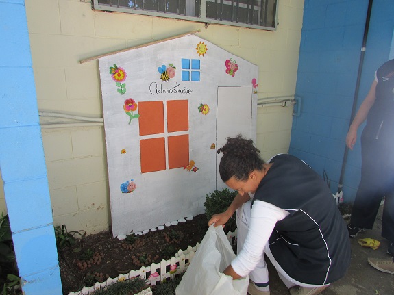 Profissional mexe em uma sacola de plástico branco. Ao fundo, a ilustração de uma casa com flores, janelas e borboletas. Ao centro da casa, janelas laranjas e escrito: Administração.