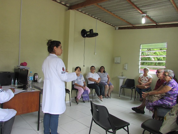 Nutricionista de jaleco branco com o logo da Secretaria Municipal da Saúde de São Paulo conversa com pacientes que estão sentados em círculo.
