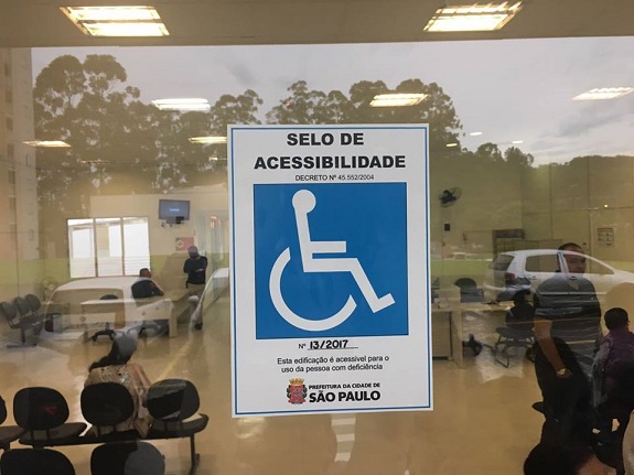 Colado na janela de vidro o selo de acessibilidade. Acima do selo o número do decreto, 45.552/2004. Abaixo, está escrito: Número 13/2017. Esta edificação é acessível para o uso da pessoa com deficiência. Na parte inferior, o logotipo da Prefeitura de São Paulo
