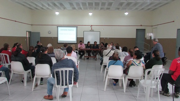 Em um auditório com pouca luz, cadeiras brancas de plástico são ocupadas por participantes. Ao fundo, uma mesa de madeira e 5 cadeiras atrás onde pessoas estão sentadas. Um projetor exibe imagens em uma tela branca.