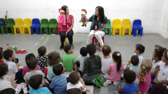 Crianças sentadas no chão assistindo ao teatro de fantoches. Ao fundo, cadeiras pequenas coloridas