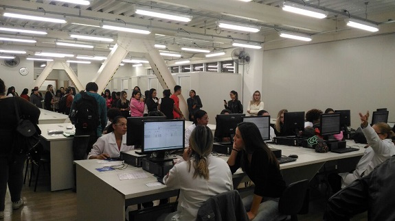 No laboratório de informática, profissionais e participantes fazem a intergração. Ao fundo, duas pilastras em forma de A preenchem o espaço da sala.