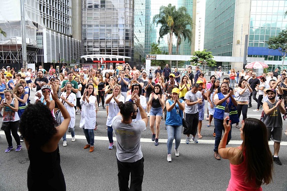 Participantes batem palmas em dança aeróbica na Avenida paulista. Ao fundo, prédios espelhados e um coqueiro.