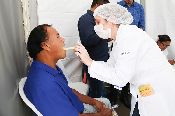 Profissional com jaleco branco, touca e luvas analisa a saúde bucal do paciente com um palito.