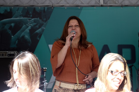 Rosana Ferro com uma camisa marrom, fala ao microfone. Ao fundo o banner da Virada da Saúde