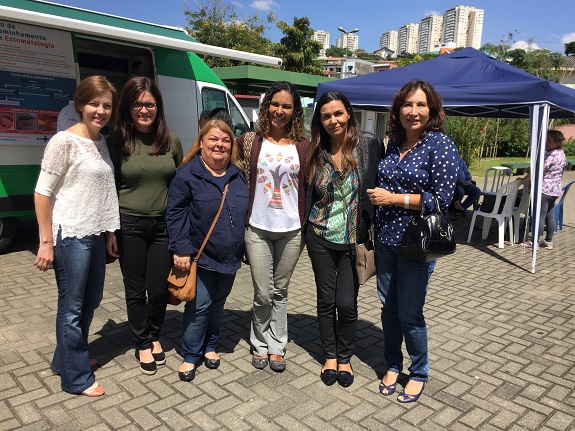 Coordenadoras posam no lado externo. Ao fundo, uma tenda azul e uma van verde e branca.