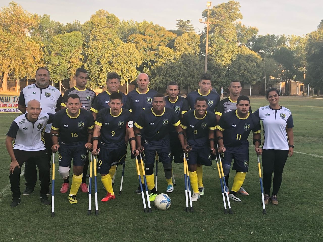 Descrição da Imagem: todos os jogadores da seleção de amputados reunidos. Eles estão em fileiras para foto. Usam muletas. Estão ao redor de um campo de futebol com gramado e árvores.