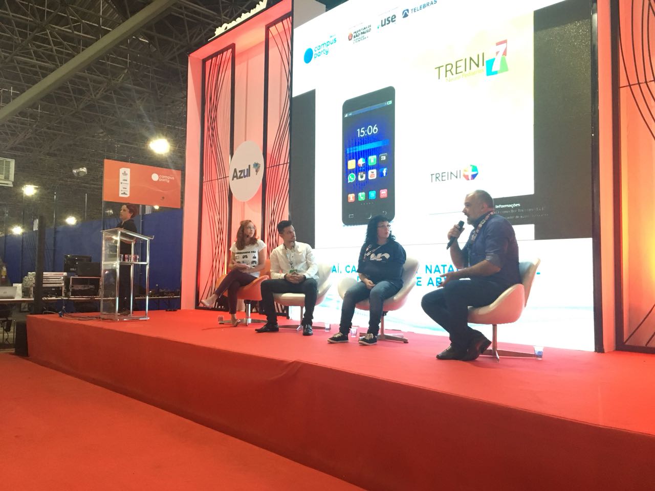  mediadora e palestrantes no palco. Eles estão sentados. Lado esquerdo do palco, a intérprete de Libras. Atrás, um telão com a imagem de um celular.