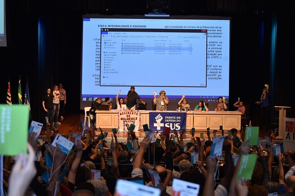 Em um auditório pouco ilumidado, participantes erguem seus crachás para votar. Ao fundo, um palco com uma mesas retangular e uma tela projetando arquivos. Ainda no palco, a bandeira do SUS e movimento das mulheres.