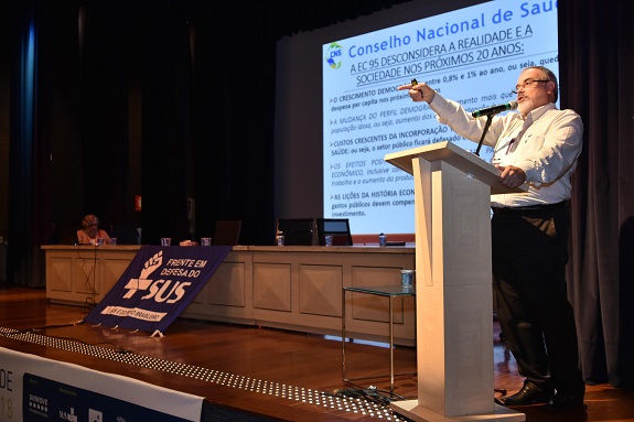 No palco, à direita, o palestrante, atrás de um pulpito, veste camisa social branca e aponta o dedo para a plateia. Ao fundo uma tela projeta um texto com o título: Conselho Nacional de Saúde.à esquerda, ainda no palco, pessoas à mesa e em frente a bandeira do SUS