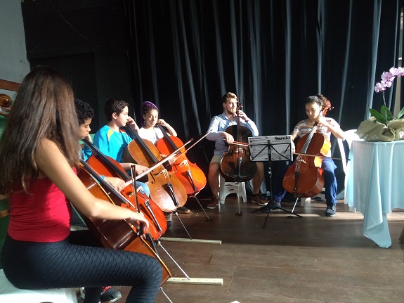 Em um palco, cinco pessoas, entre elas 4 adultos e duas crianças, tocam violoncelo. Ao fundo, uma cortina preta. À direita, uma mesa com toalha branca e um vaso de orquídea decoram o ambiente.