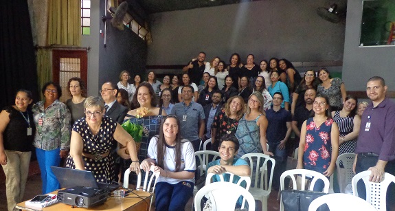 Em um salão participantes posam para a foto. À frente, uma mesa com um retoprojetor e um notebook.