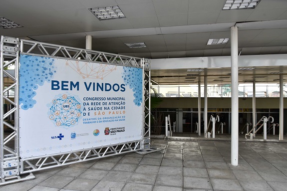 Painel na entrada do Centro de Convenções do Anhembi em um fundo branco com os seguintes dizeres em azul marinho: Bem-vindos ao Congresso Municipal da Rede de Atenção à Saúde na Cidade de São Paulo - Desafios da Organização do Trabalho e da Educação na Saúde - Desafios da Organização do Trabalho e da Educação na Saúde