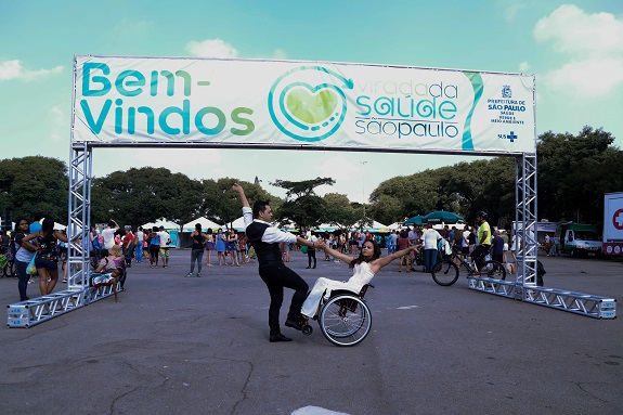 Uma placa de entrada branca, com letras em azul e verde e logo da Virada da saúde diz: Bem- Vindos; Virada da Saúde são Paulo. Ao centro, um casal em posição de dança, com braços esticados. A moça está em uma cadeira de rodas. Ao fundo, tendas e árvores verdes.