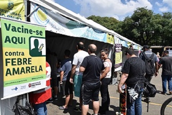 Tendas de saúde e pessoas circulando. À esquerda, um banner verde escrito: Vacine-se aqui contra a Febre amarela.